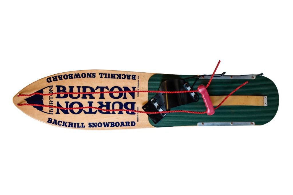 A vintage snowboard that has a narrow tip, rudimentary bindings, and a rope to hold to control it.