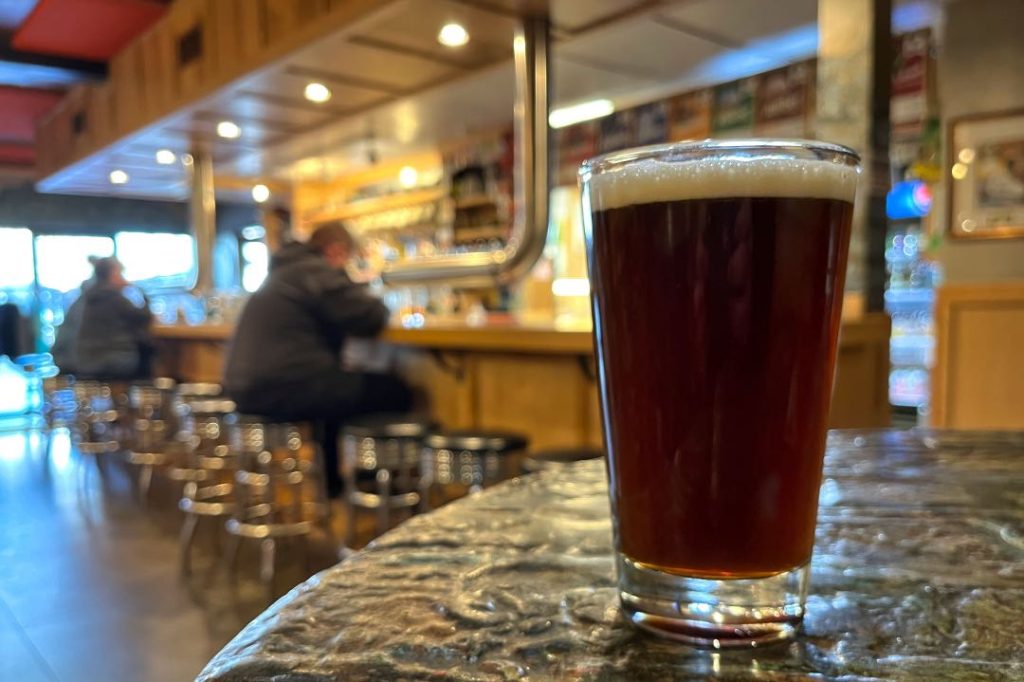 A pint glass of dark brown beer on table, in the background is blurred scene of bar patrons sitting on stools at the bar.